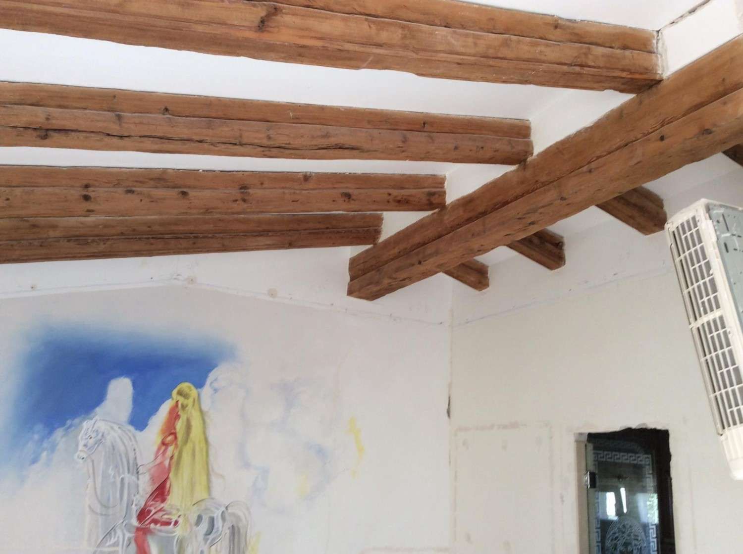 Un techo blanco con vigas rústicas de madera a la vista y un mural de pared pintado con formas abstractas azules y amarillas.