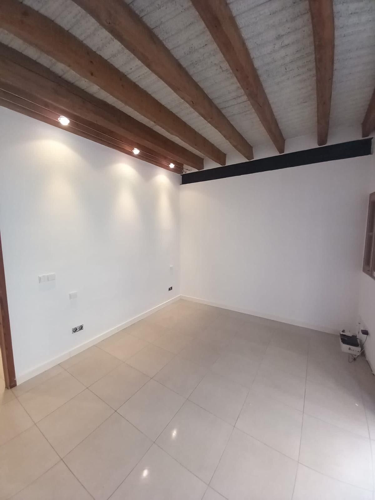 Habitación vacía con paredes blancas, suelo de baldosas y vigas de madera en el techo con iluminación empotrada.