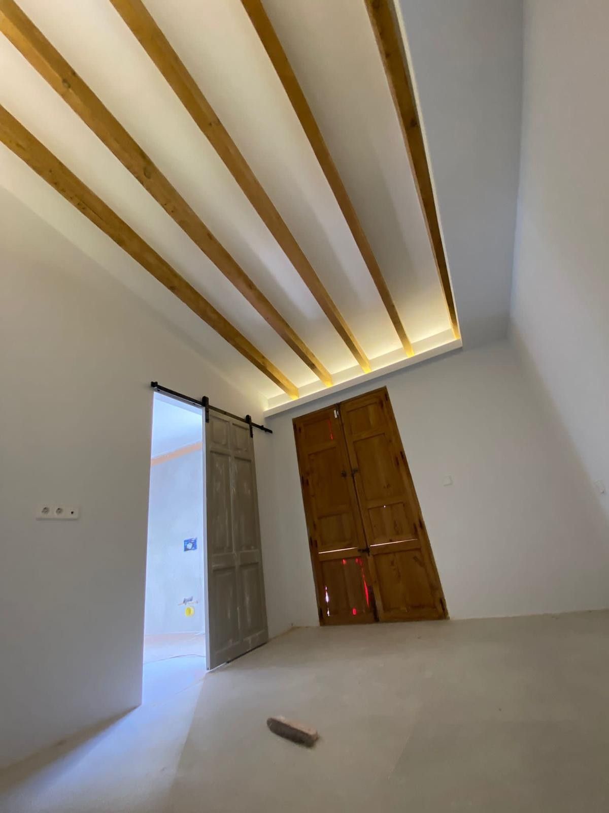 Una habitación con paredes blancas, piso de concreto, vigas de madera en el techo, una puerta corrediza estilo granero.