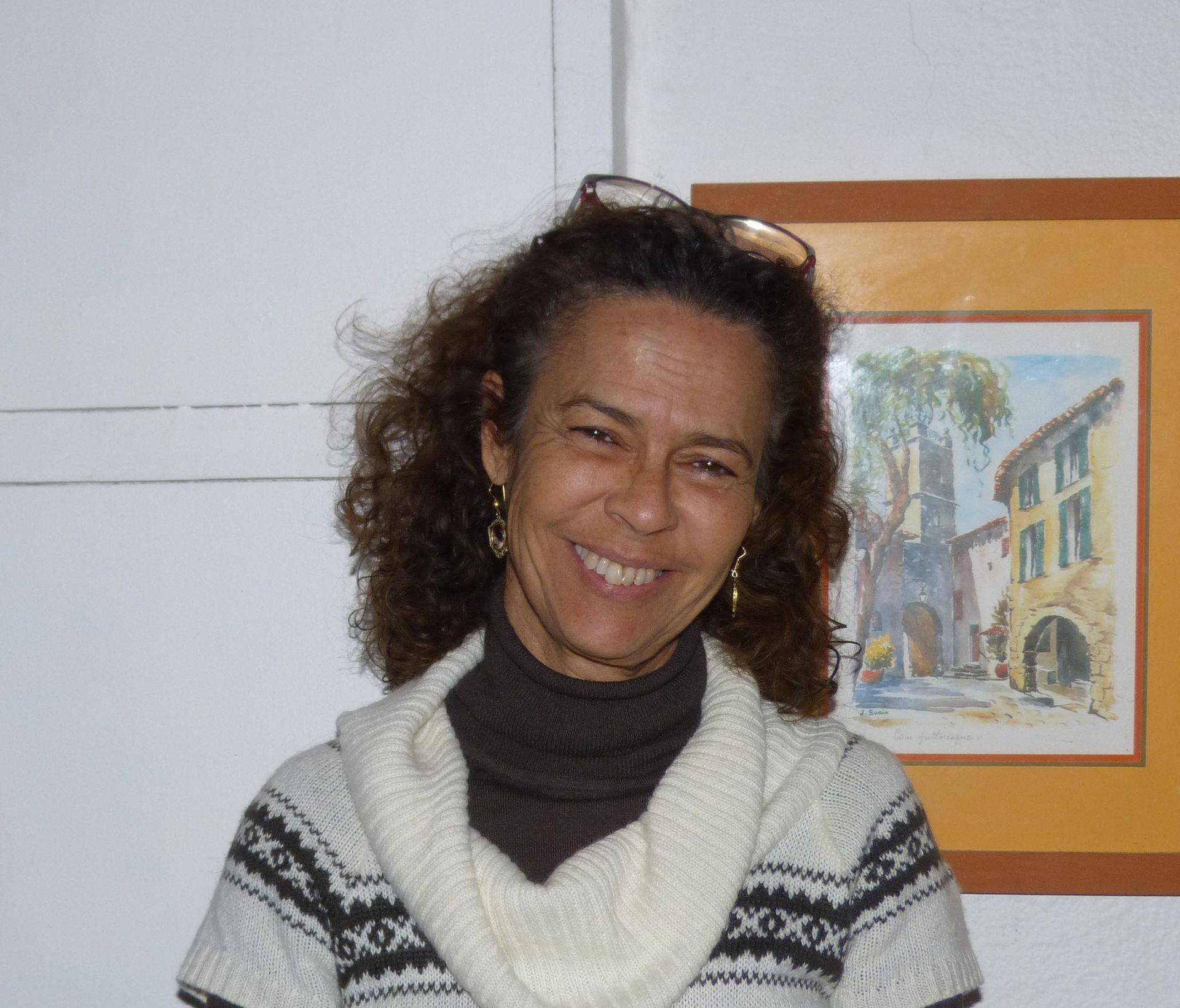 Femme aux cheveux bouclés, souriante, portant un pull, debout devant un tableau.