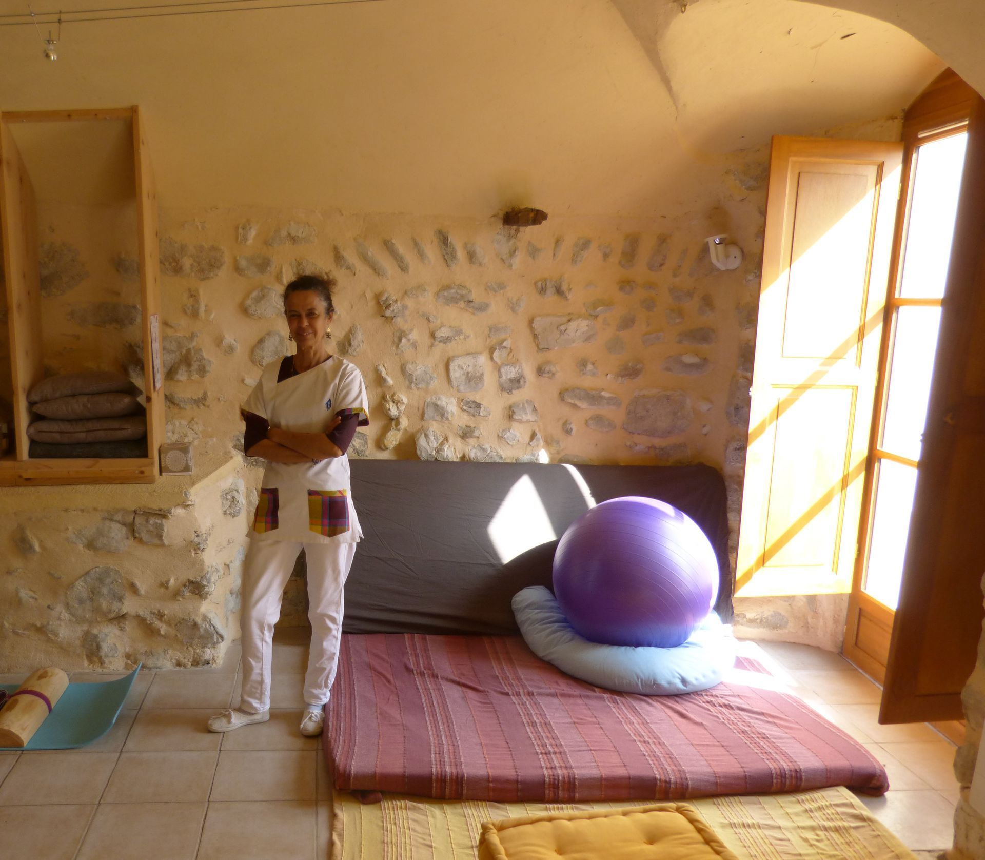 Une femme en uniforme blanc se tient dans une pièce aux murs de pierre avec un lit, un ballon d'exercice et une fenêtre ouverte.