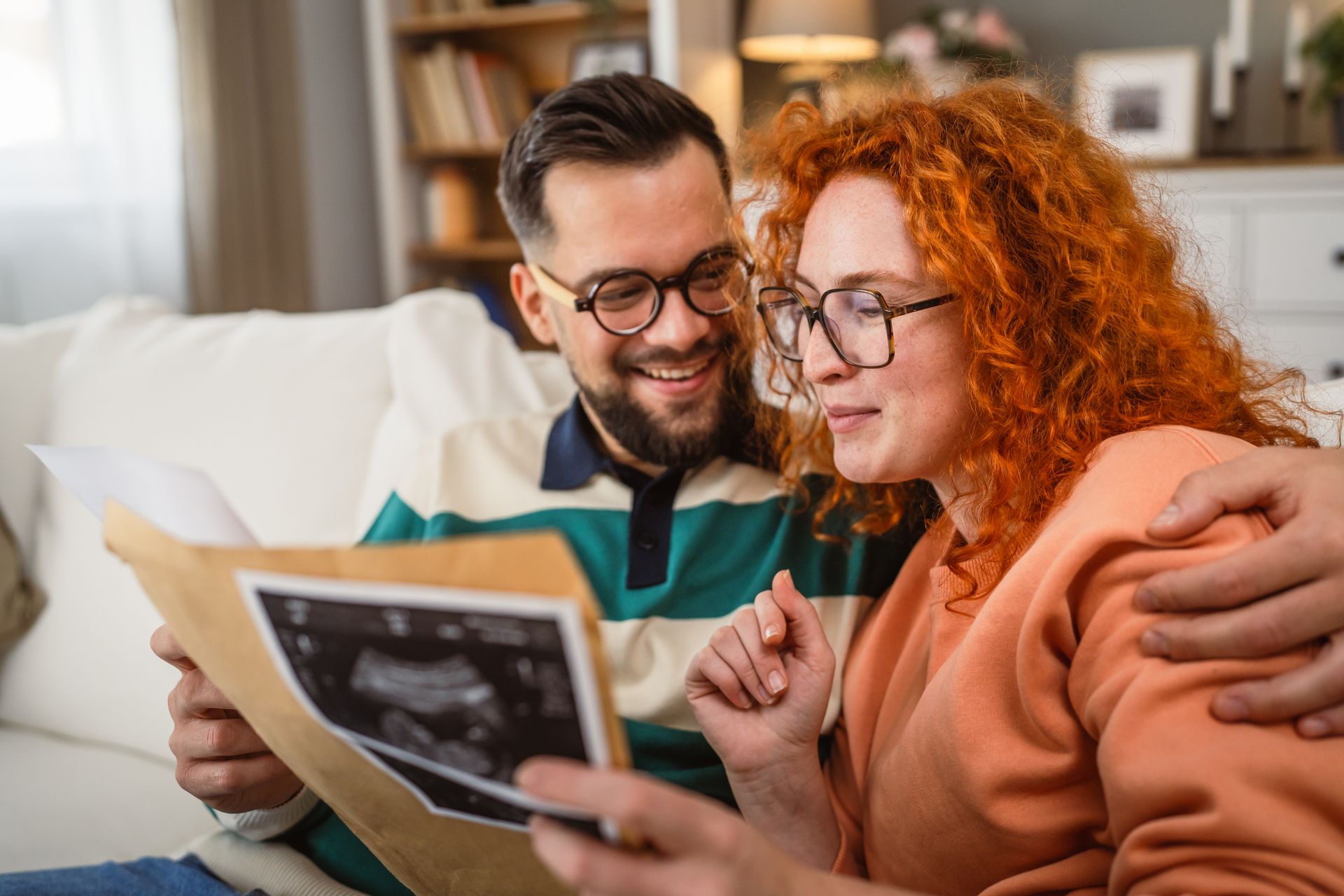 Un couple sourit en regardant une image échographique, assis sur un canapé dans un salon.