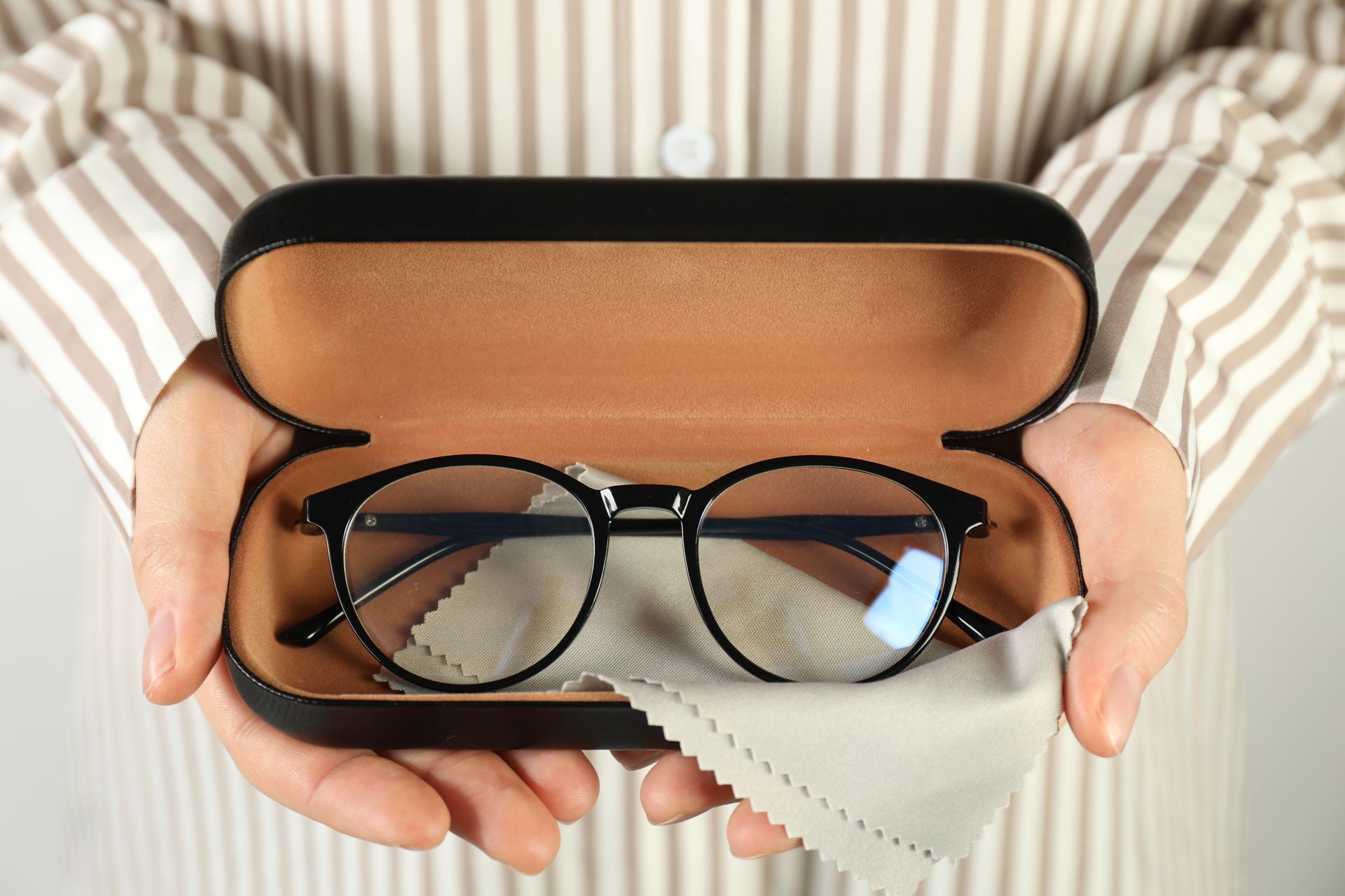 Des mains tenant un étui à lunettes ouvert contenant des lunettes à monture noire et une lingette de nettoyage