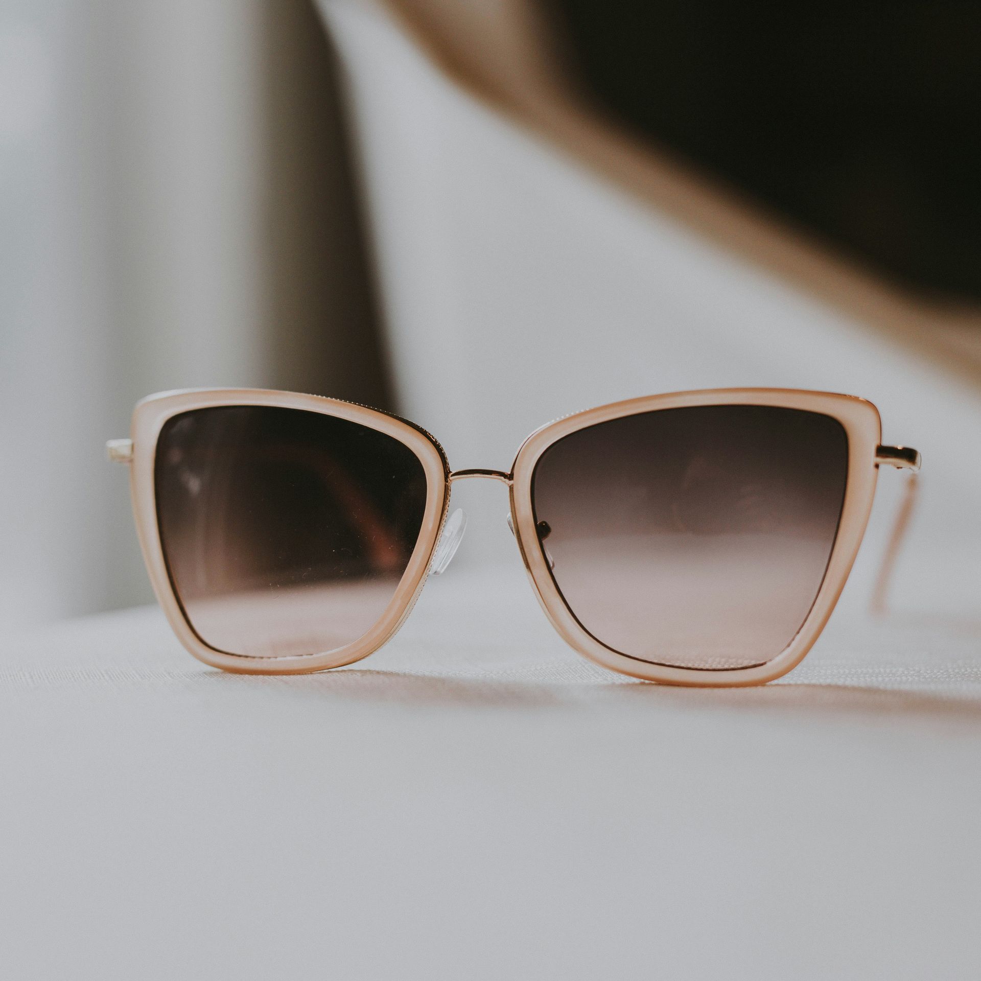 Lunettes de soleil œil-de-chat en or rose posées sur une surface blanche, avec un arrière-plan flou et doux