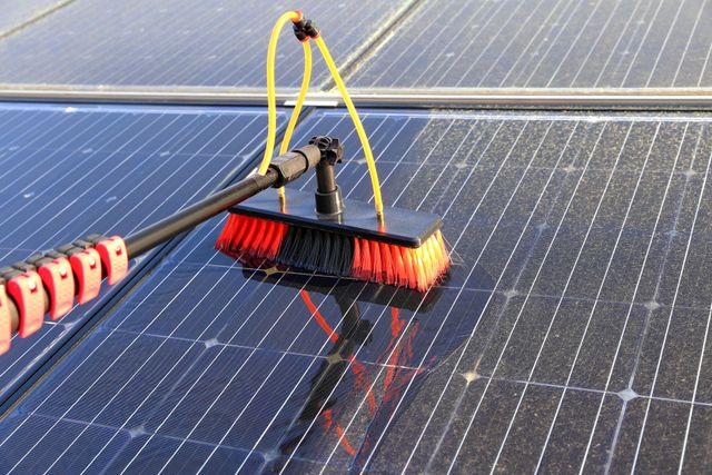 Brosse de nettoyage de panneaux solaires à poils rouges sur un toit, pulvérisant de l'eau sur des panneaux bleu foncé