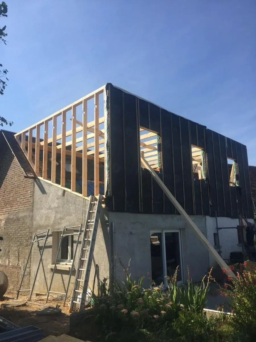 Maison en rénovation avec charpente apparente en bois et panneaux extérieurs sombres se détachant sur un ciel bleu.