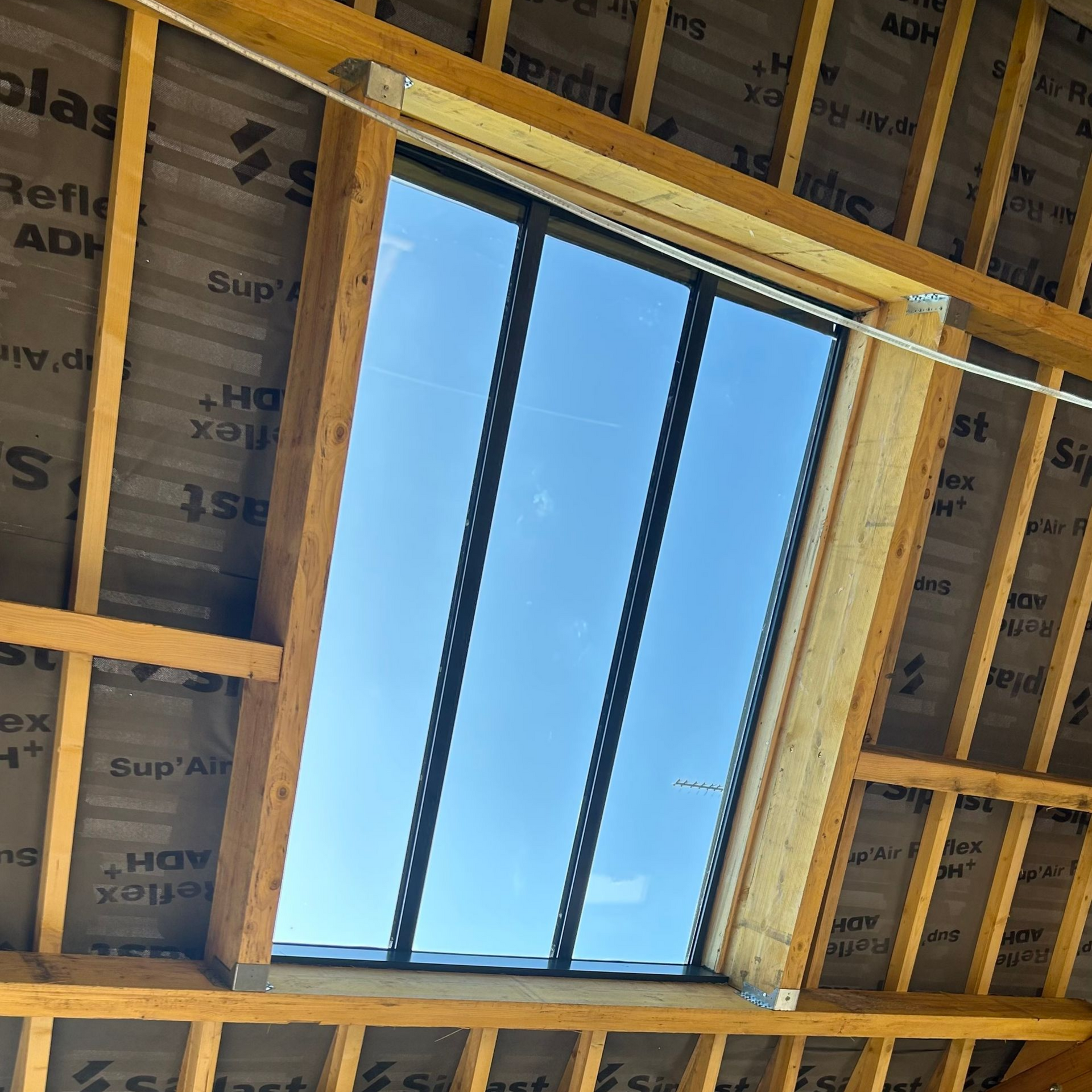 Puits de lumière dans une charpente en bois, l'isolation étant apparente tout autour, laissant entrevoir le ciel bleu à l'extérieur.