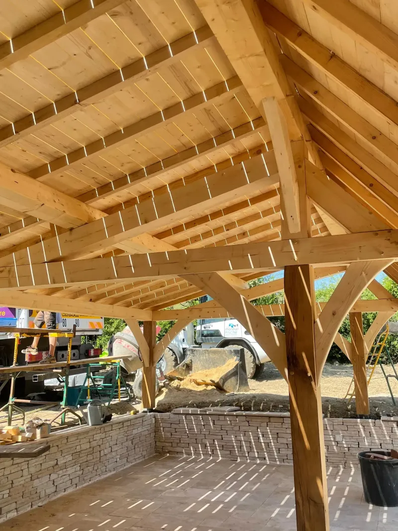 Pavillon en bois aux poutres apparentes et aux murs en pierre, donnant sur un chantier de construction avec des engins de chantier en arrière-plan.