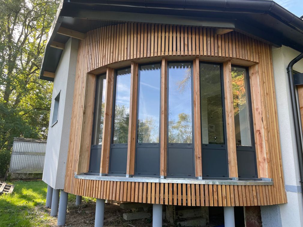 Fenêtre en baie cintrée en bois à lattes verticales sur la façade d'une maison, encadrée par des arbres et de la verdure.
