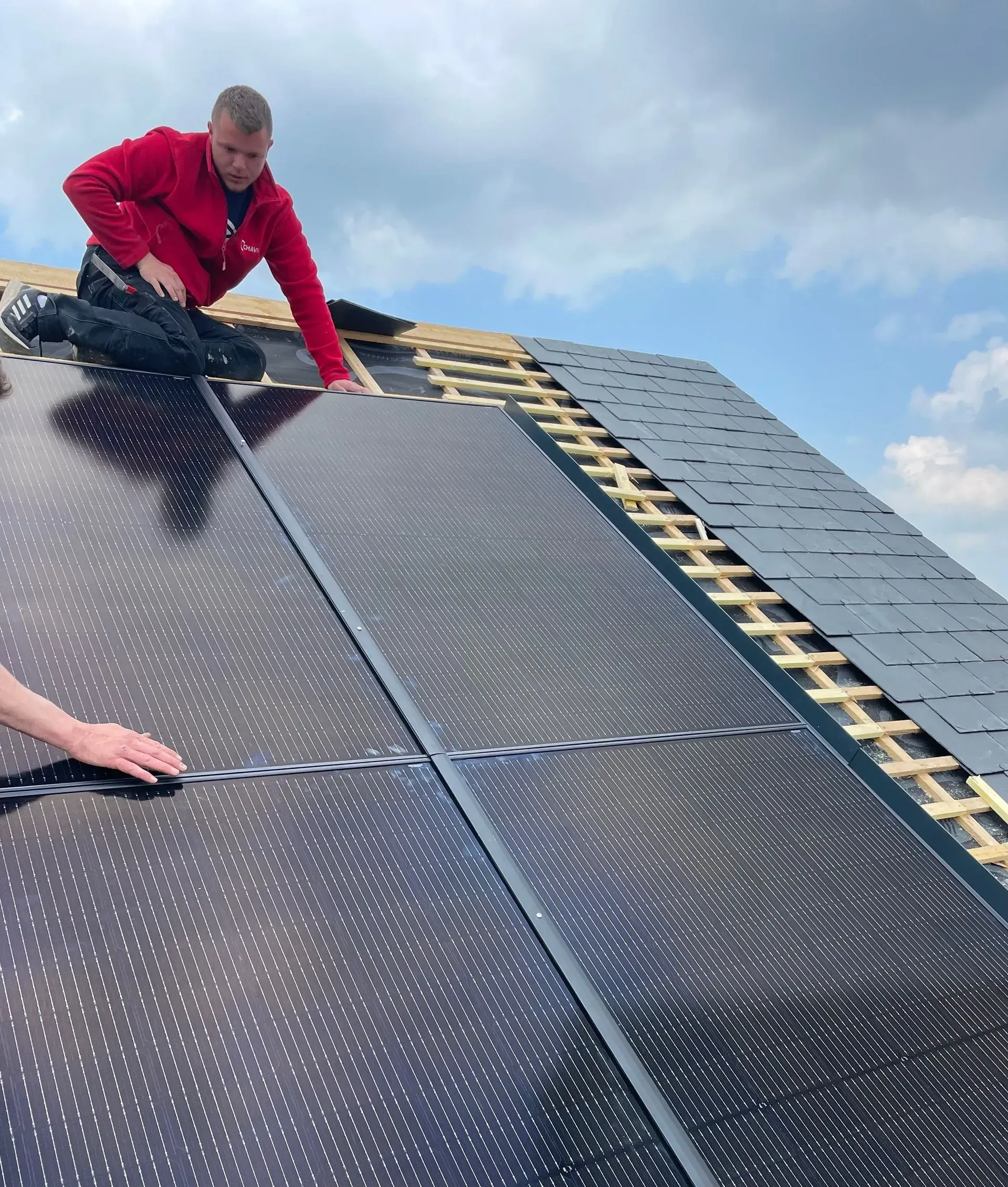 Ouvrier installant des panneaux solaires sur un toit sous un ciel nuageux.
