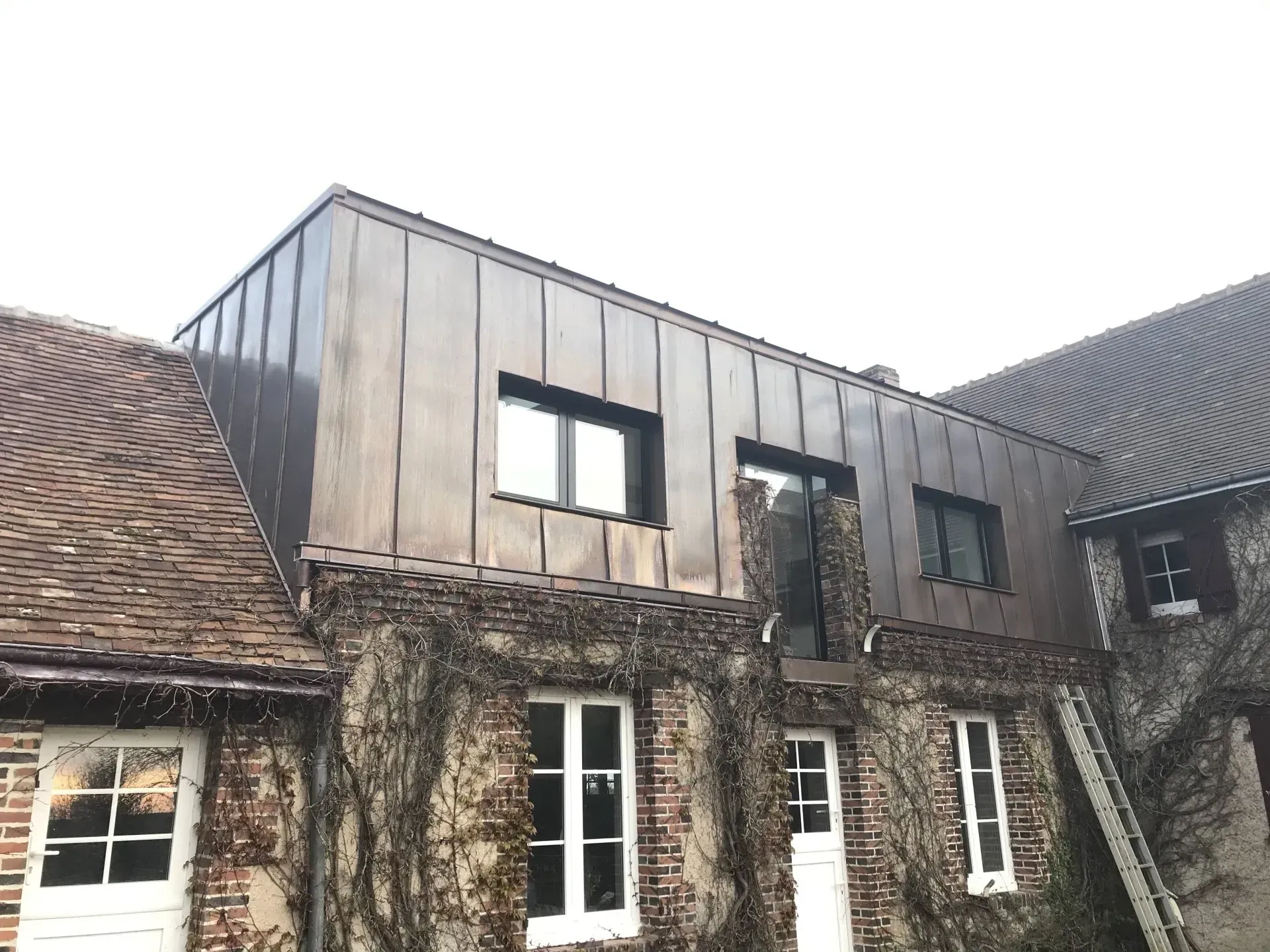 Bâtiment en pierre patinée avec une façade supérieure en métal gris et plusieurs fenêtres.