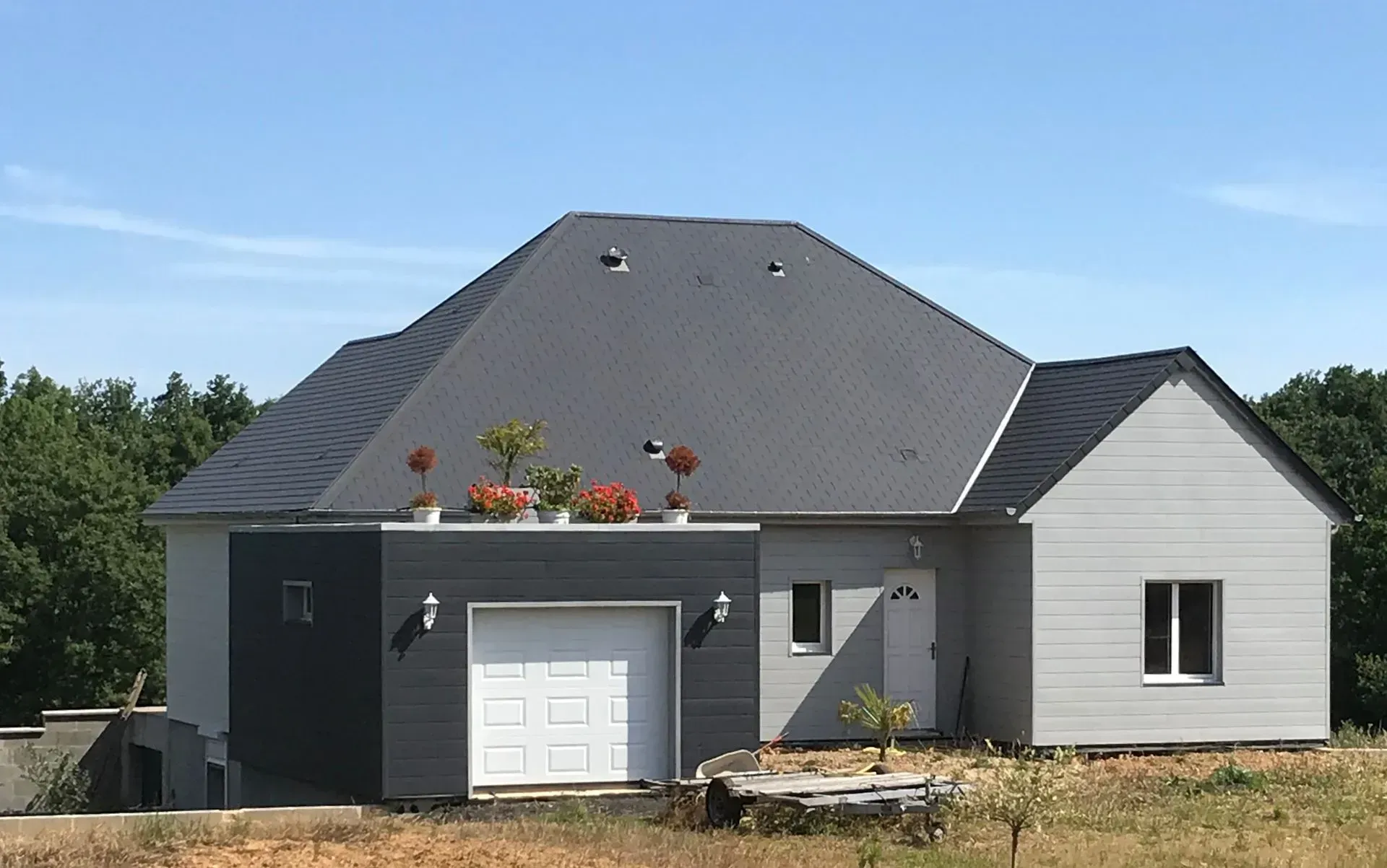 Maison moderne avec toit gris foncé, bardage gris clair et garage sous un ciel bleu azur.