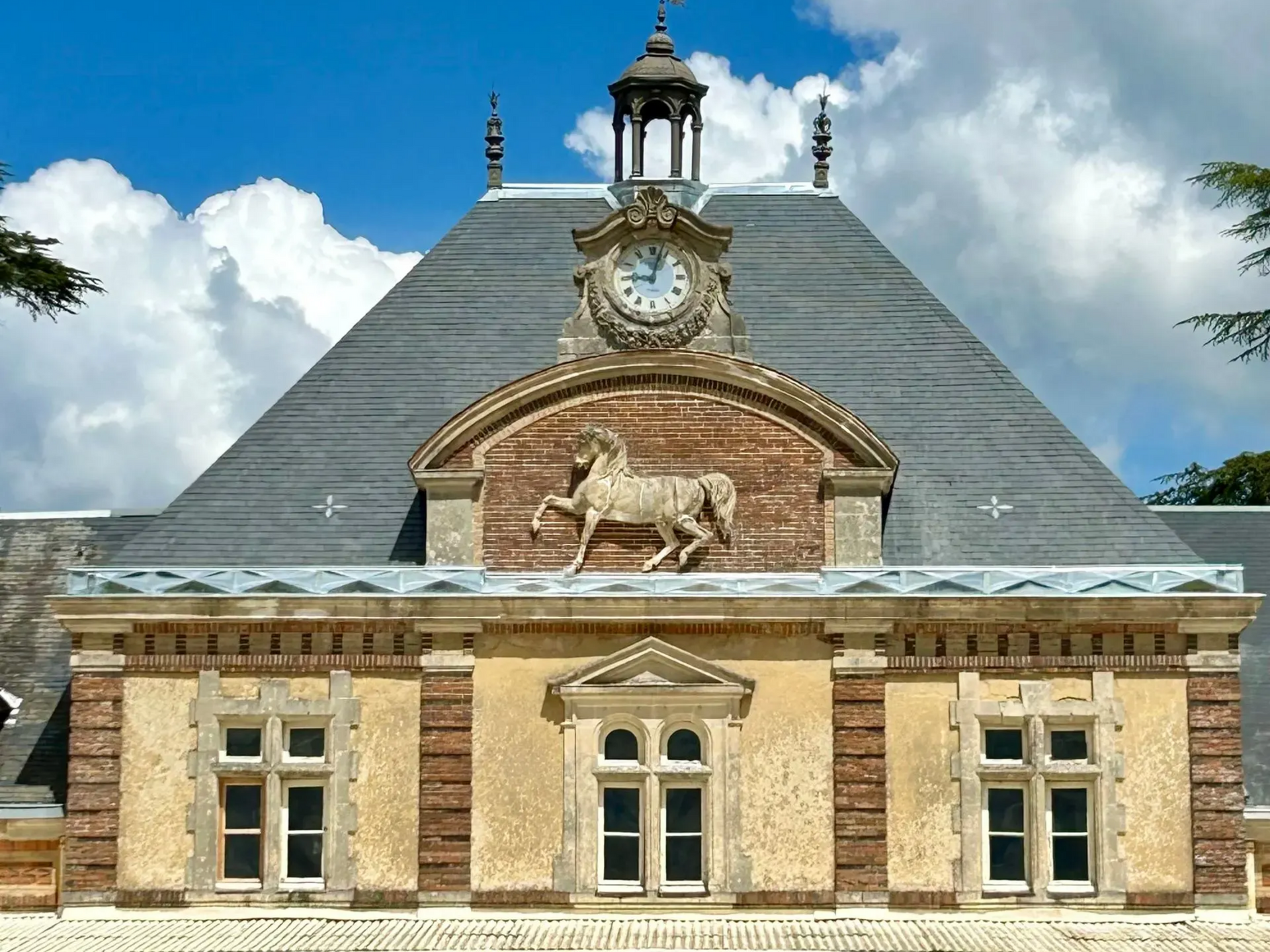 Façade historique en briques avec une tour d'horloge, un toit en ardoises et un relief animalier sous un ciel bleu