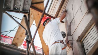 Un ouvrier du bâtiment, casqué de rouge, répare la façade d'un immeuble sur un échafaudage.