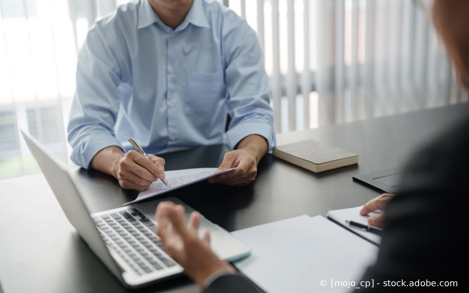 Person, die während einer Besprechung ein Dokument unterschreibt; Laptop, Notizbuch und Buch auf dem Schreibtisch.