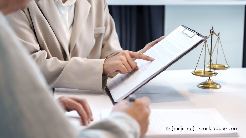 Person zeigt auf ein Dokument auf einem Klemmbrett; im Hintergrund die Waage der Gerechtigkeit.