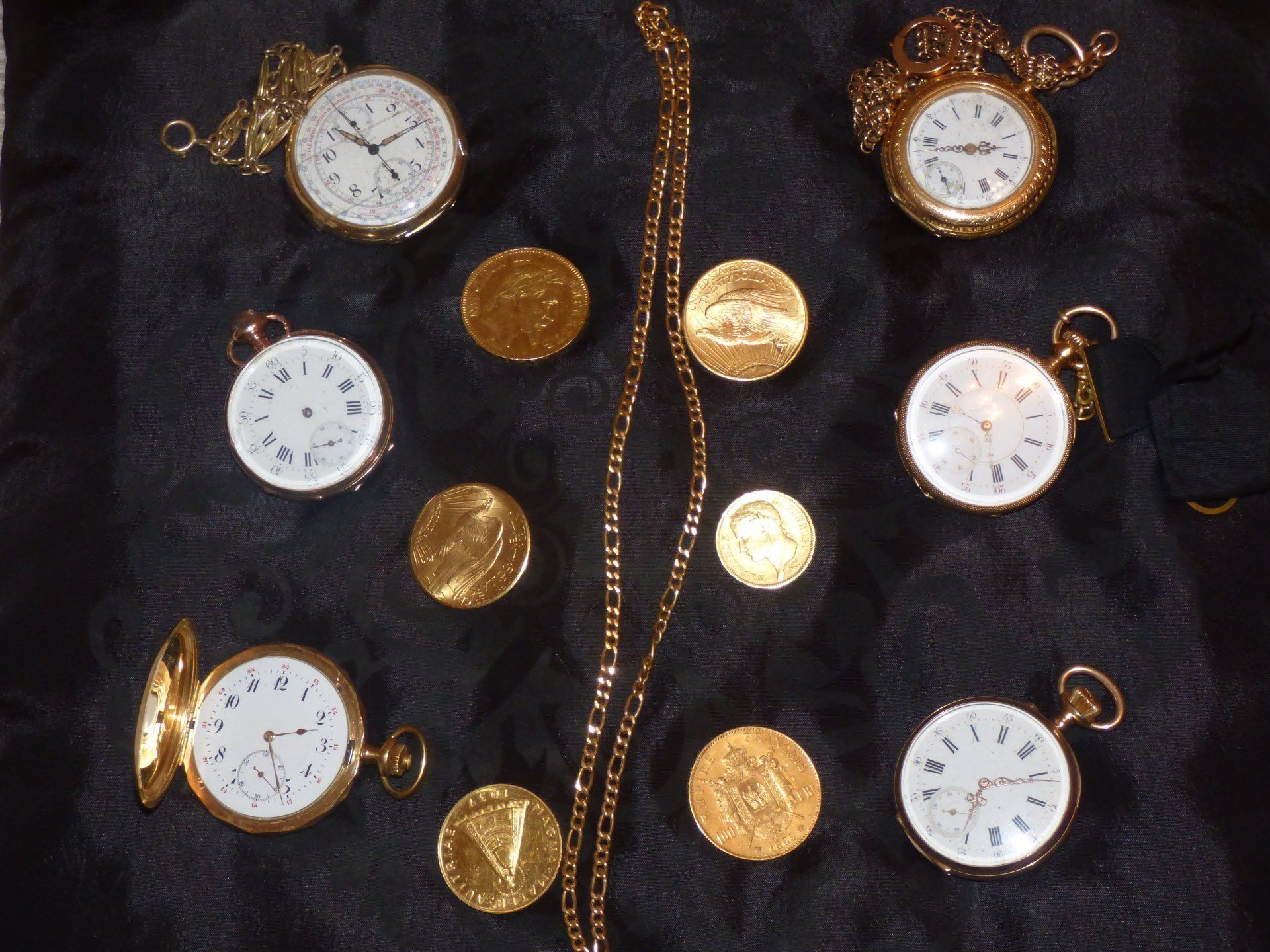 Montres de poche et pièces en or disposées sur un tissu noir. Une chaîne en or est également présente.