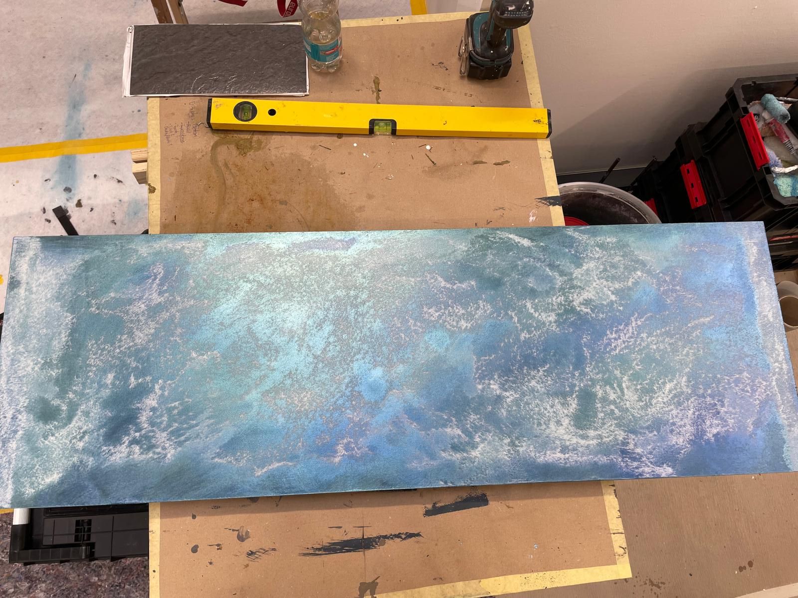 Auf einem Holztisch steht neben einer gelben Wasserwaage ein blaues Gemälde.