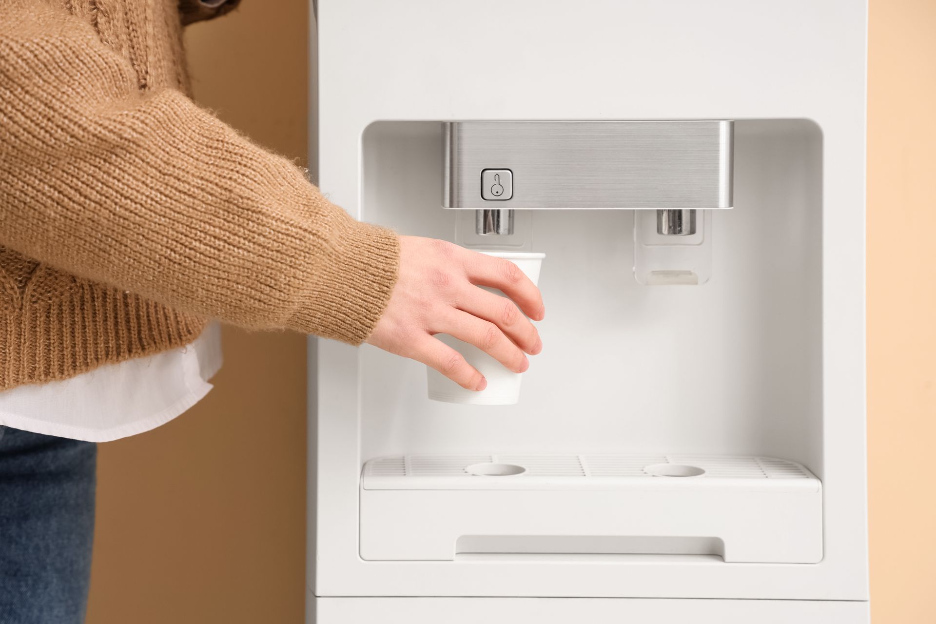 Une personne portant un pull marron tient une tasse blanche sous le bec d'une fontaine à eau blanche.