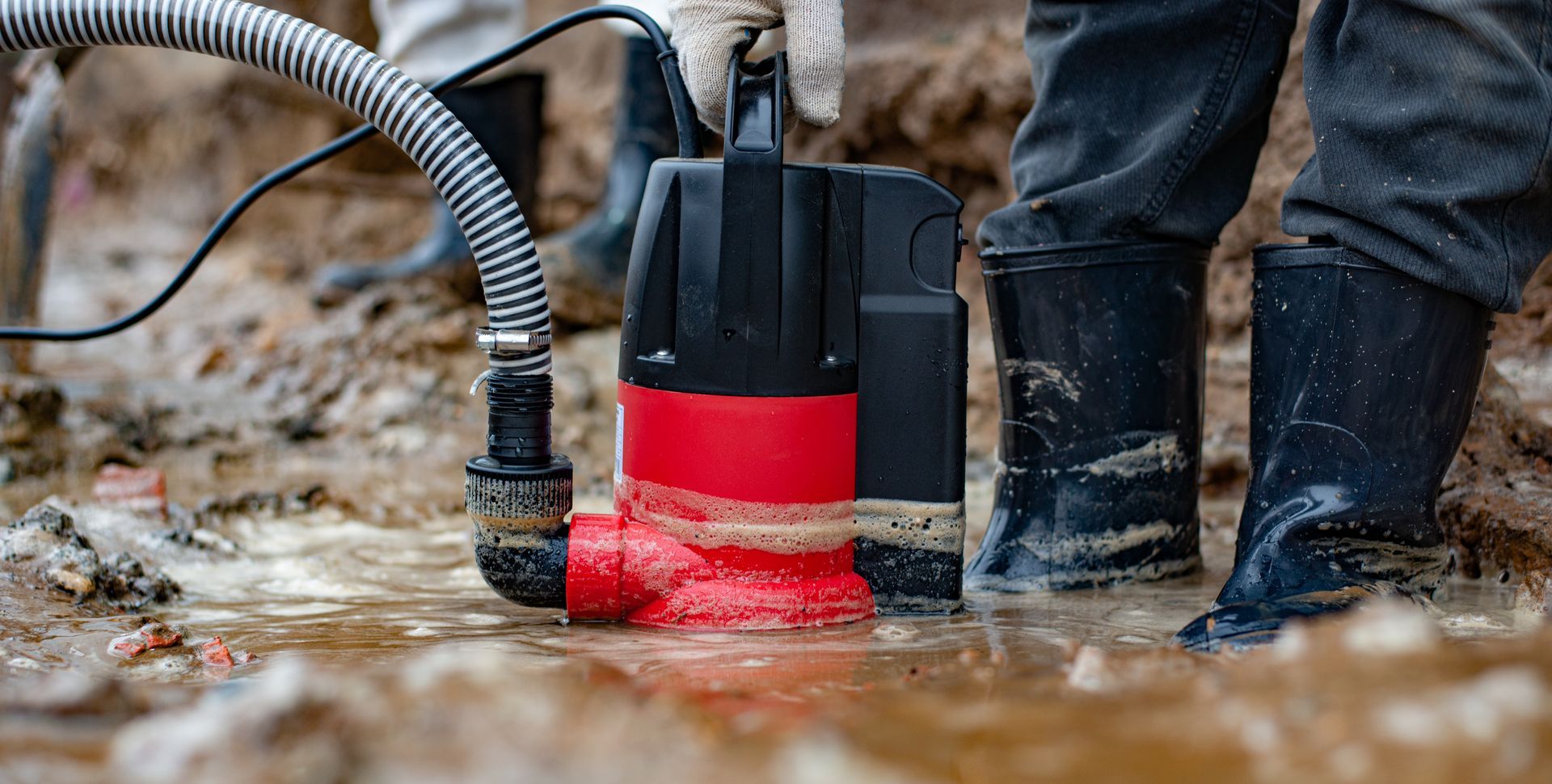 Une personne utilise une pompe à eau rouge et noire dans des eaux de crue boueuses, à côté de bottes et d'un tuyau d'arrosage.