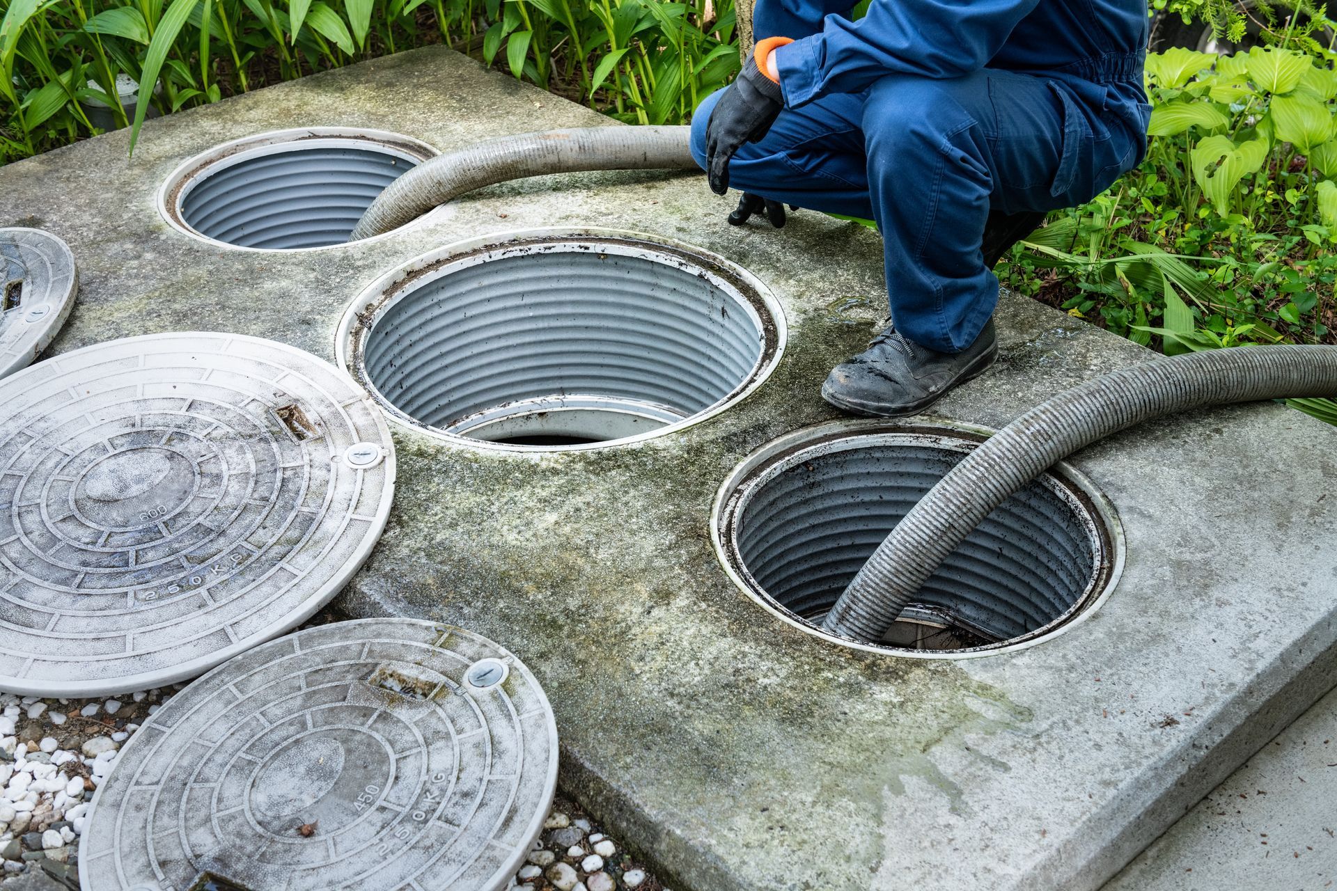 Un ouvrier inspecte des regards d'égout ouverts, couvercles relevés, sur une surface en béton à l'extérieur.