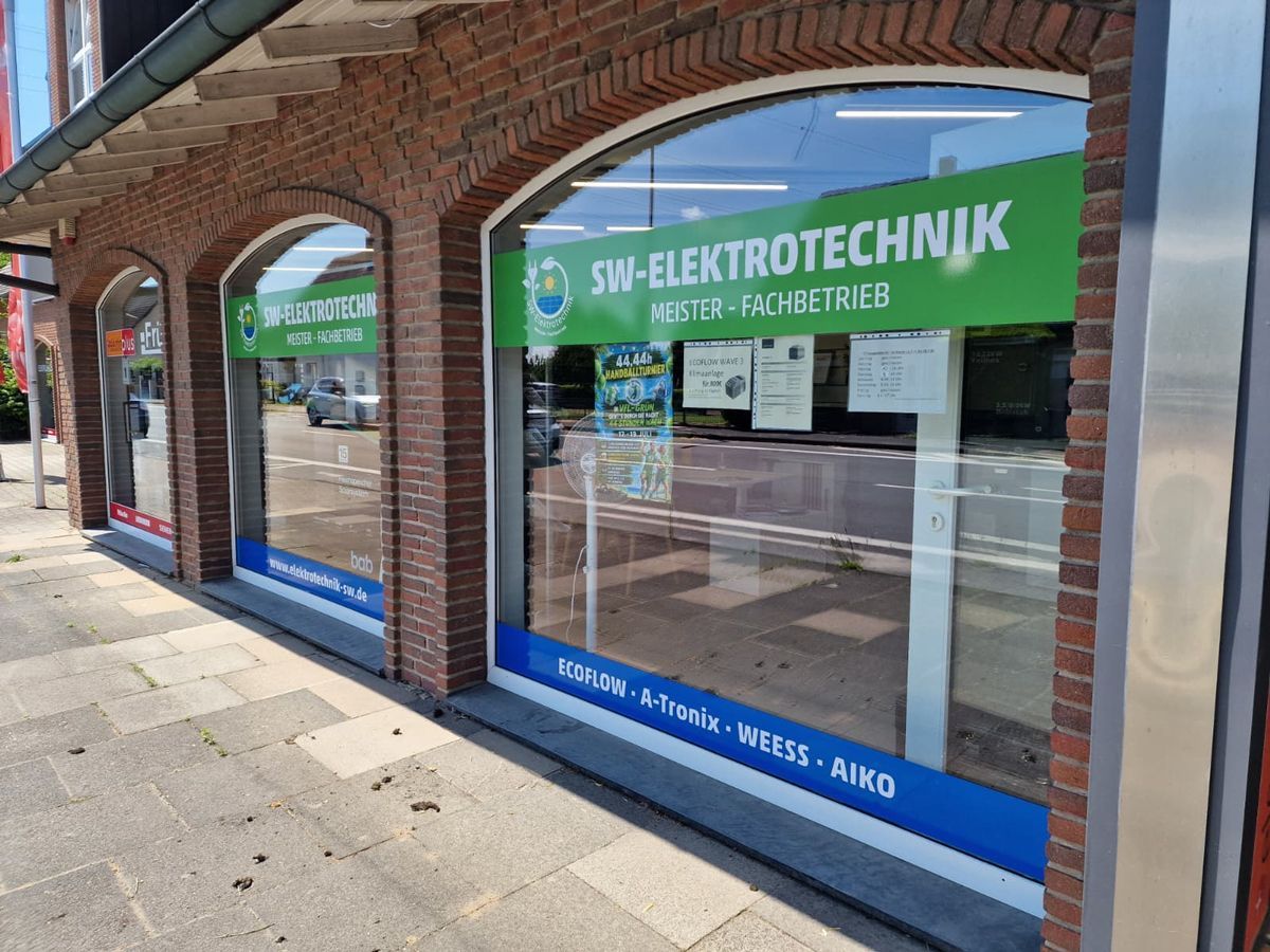 Schaufenster mit grünem „SIT-ELEKTROTECHNIK“-Schild und blauen Fensterbannern auf einem Backsteinpflasterweg.