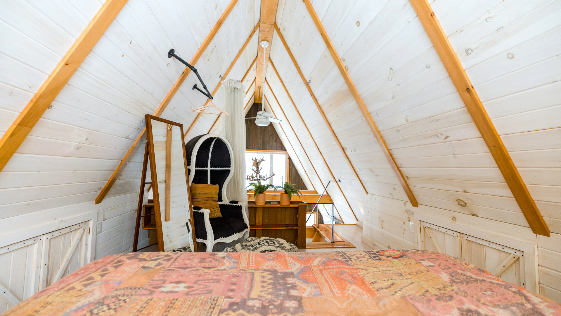 Chambre mansardée confortable avec plafond en bois blanc incliné, lit, chaise et petit bureau près d'une fenêtre.