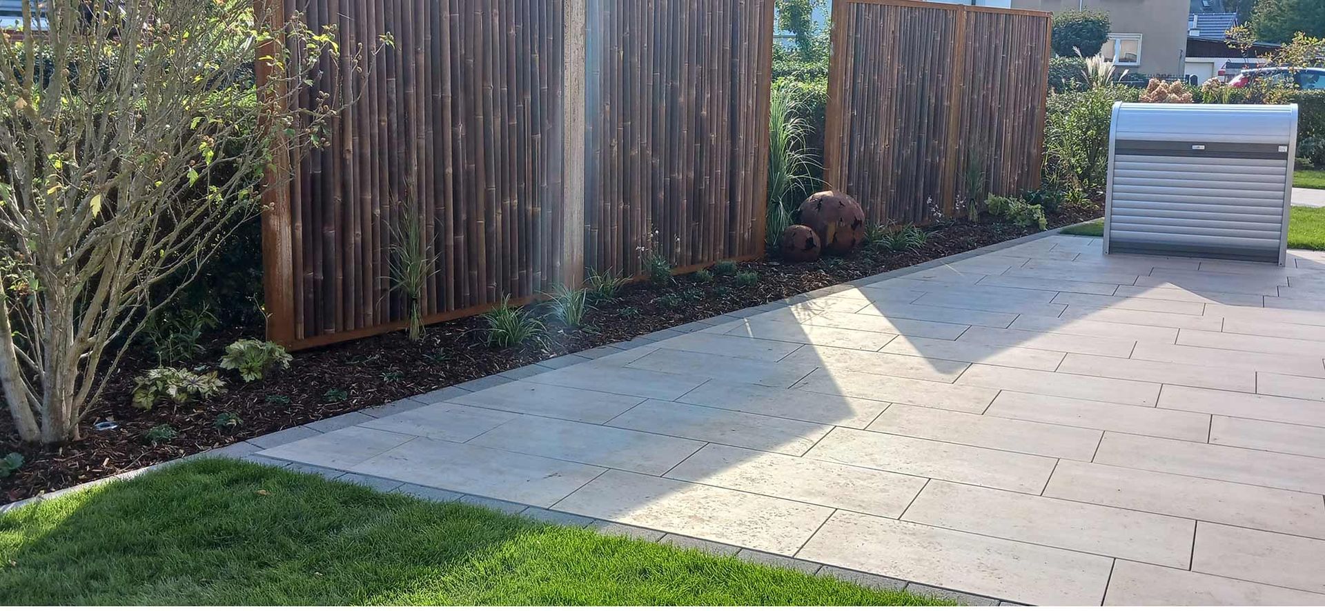 Eine Beton-Terrasse mit Holzzaun und Grünpflanzen. Im Hintergrund ist ein Briefkasten zu sehen.