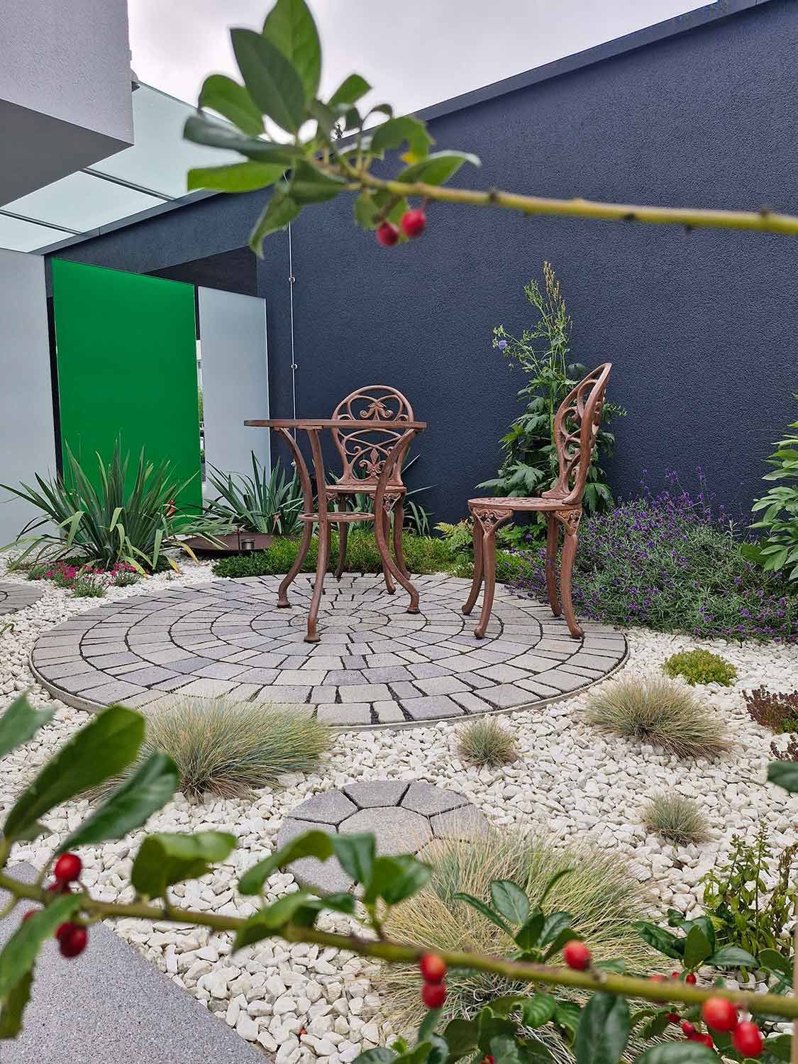 Ein kleiner Garten mit einer Ziegelterrasse, einem schmiedeeisernen Tisch und Stuhl sowie weißem Kies. Grüne und schwarze Wände.