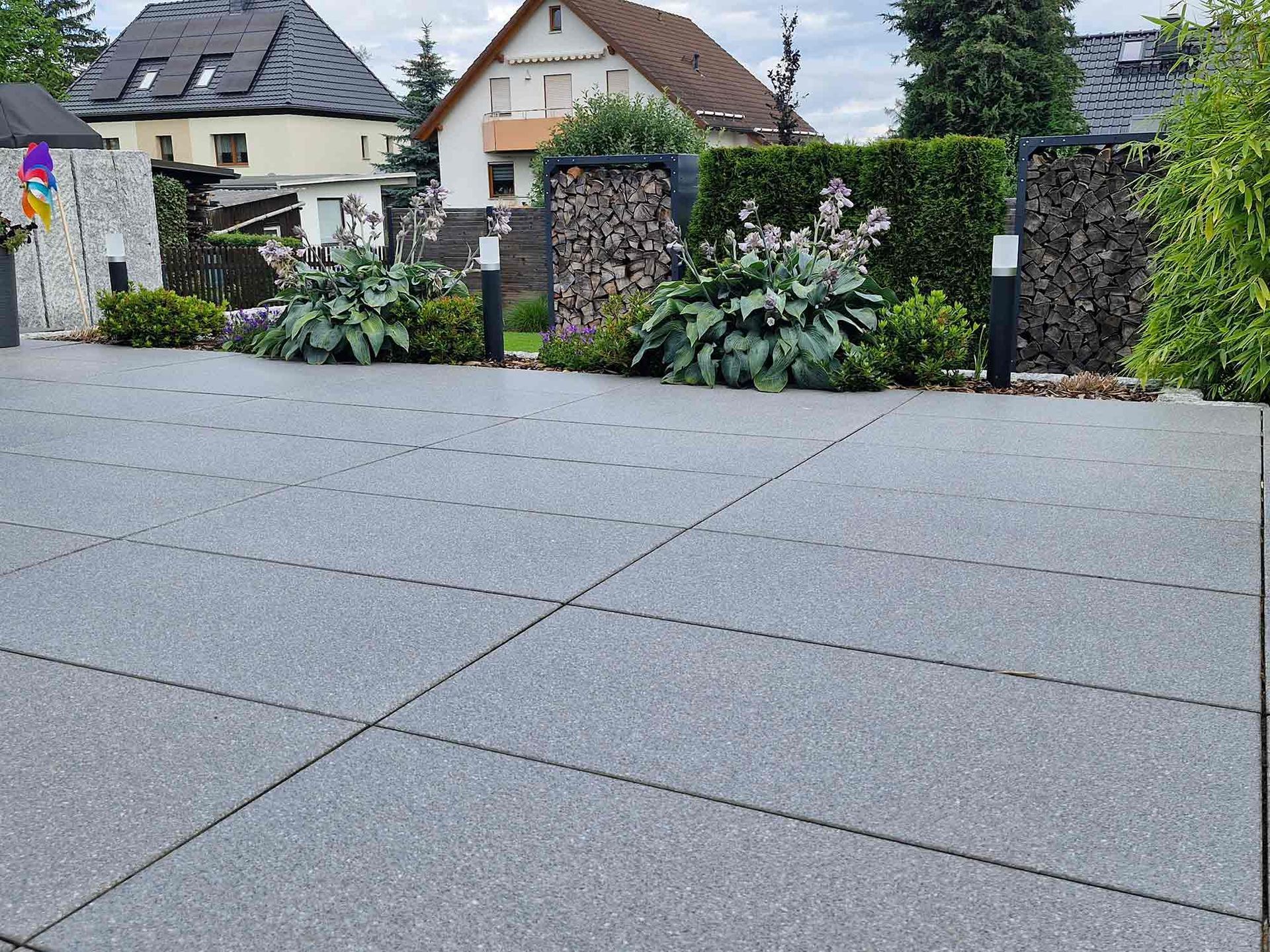 Grau gepflasterte Auffahrt mit bepflanztem Randstreifen, Steinmauer und Häusern im Hintergrund.