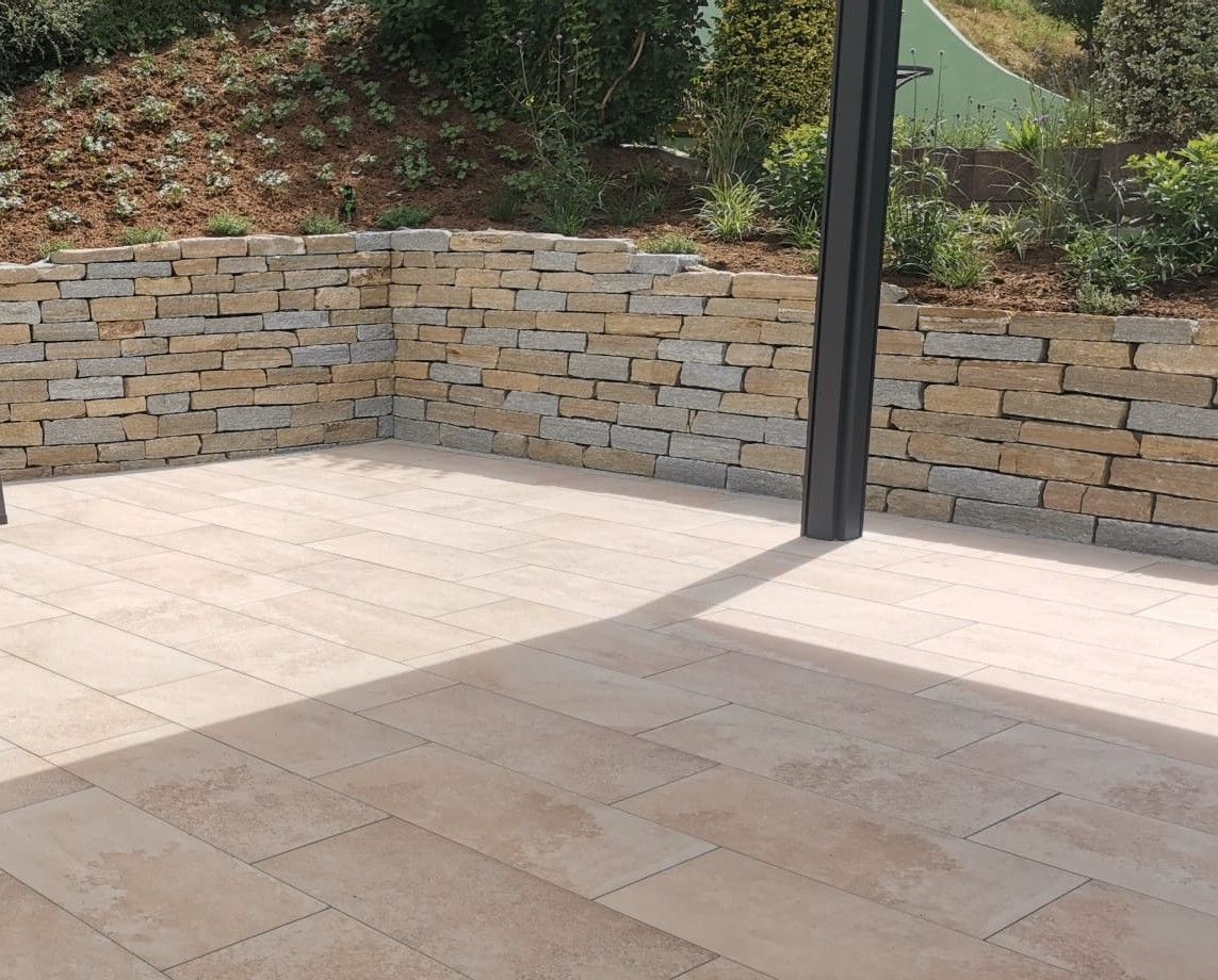 Terrasse mit beigefarbenem Fliesenboden und Steinmauer, beschattet von einer dunkelgrauen Stütze.