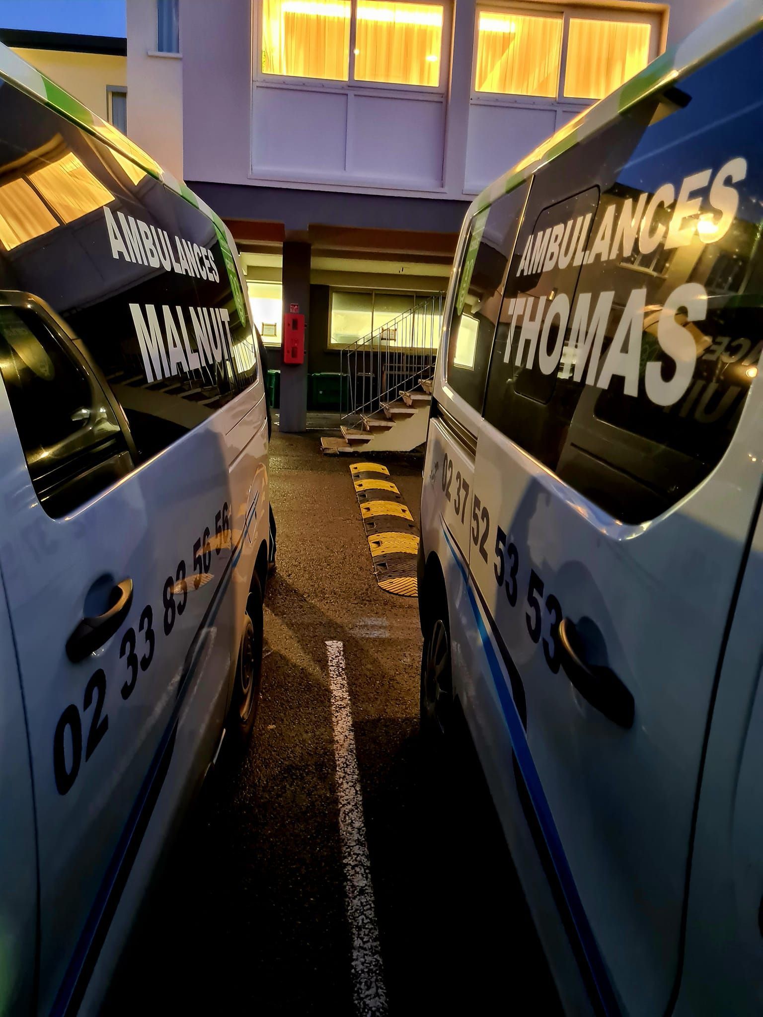 Deux ambulances, l'une avec le logo Ambulances Thomas et l'autre, le logo Ambulances Malnuit garées l'un à côté de l'autre dans un parking la nuit