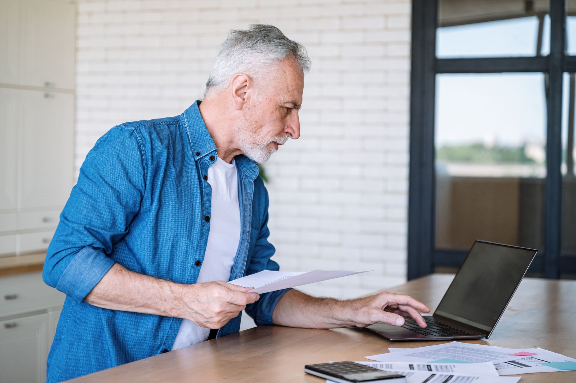 Un homme âgé tenant une feuille à la main consulte son ordinateur portable
