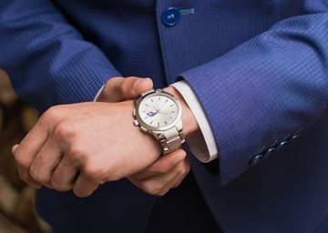 Persona vestida con un traje azul revisando un reloj de pulsera plateado en su muñeca izquierda.