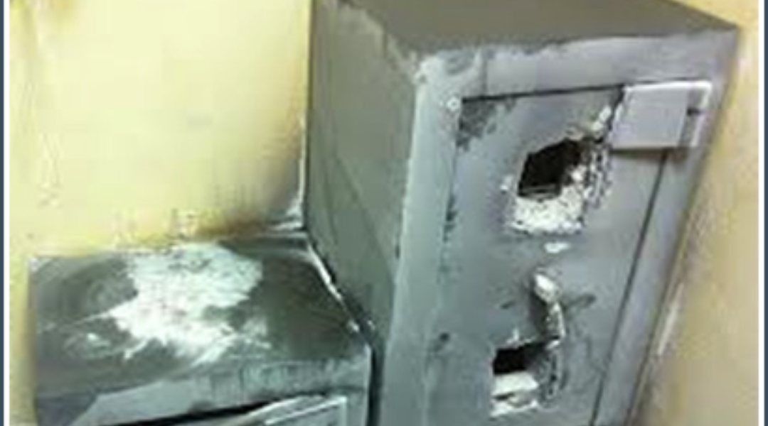 Caja fuerte gris dañada, con agujeros, apoyada contra una pared, con aspecto de haber sido forzada.