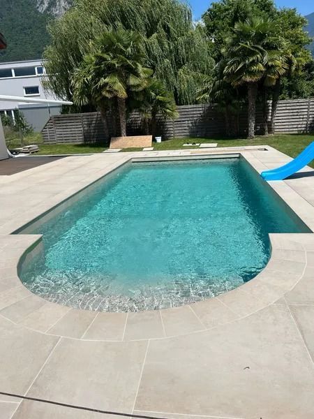 Piscine entourée de pierres claires. Palmiers et toboggan bleu à proximité.