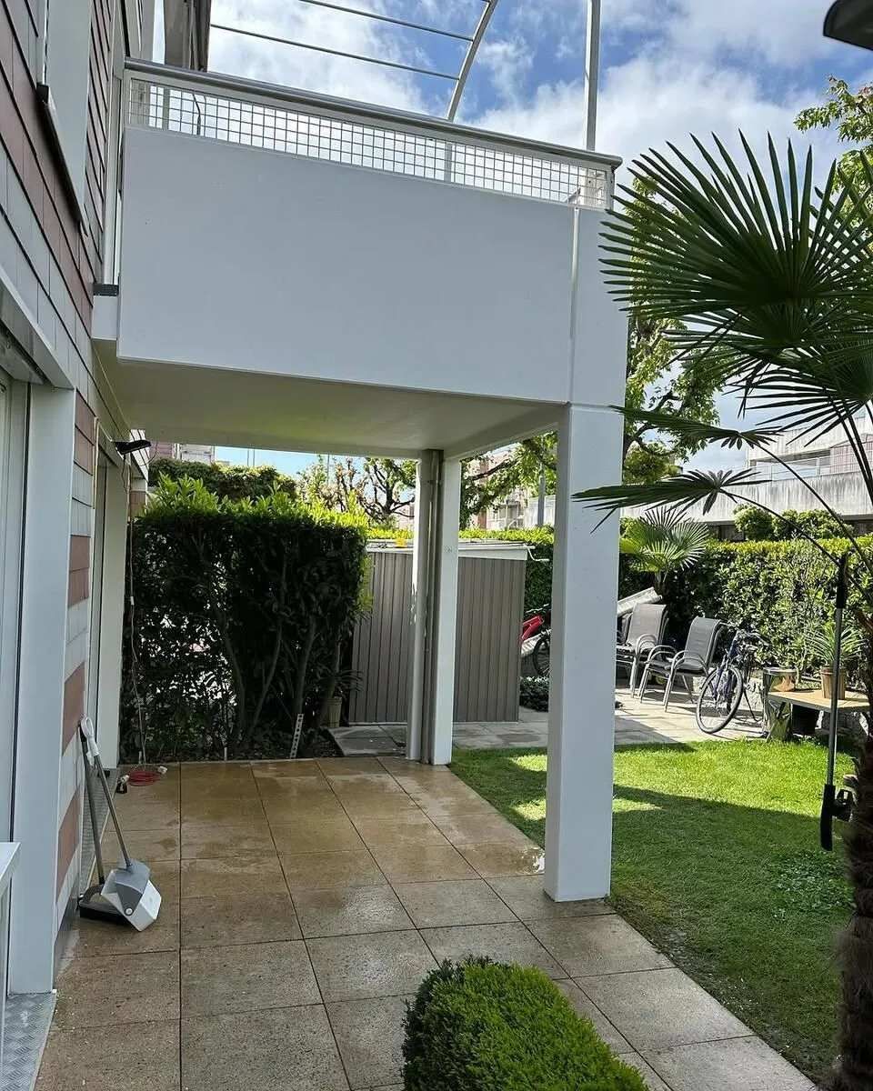 Patio couvert avec piliers blancs, à côté d'une maison avec verdure et mobilier d'extérieur.