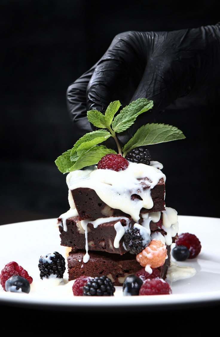 Una persona que lleva guantes negros sostiene una pila de brownies con bayas en un plato blanco.