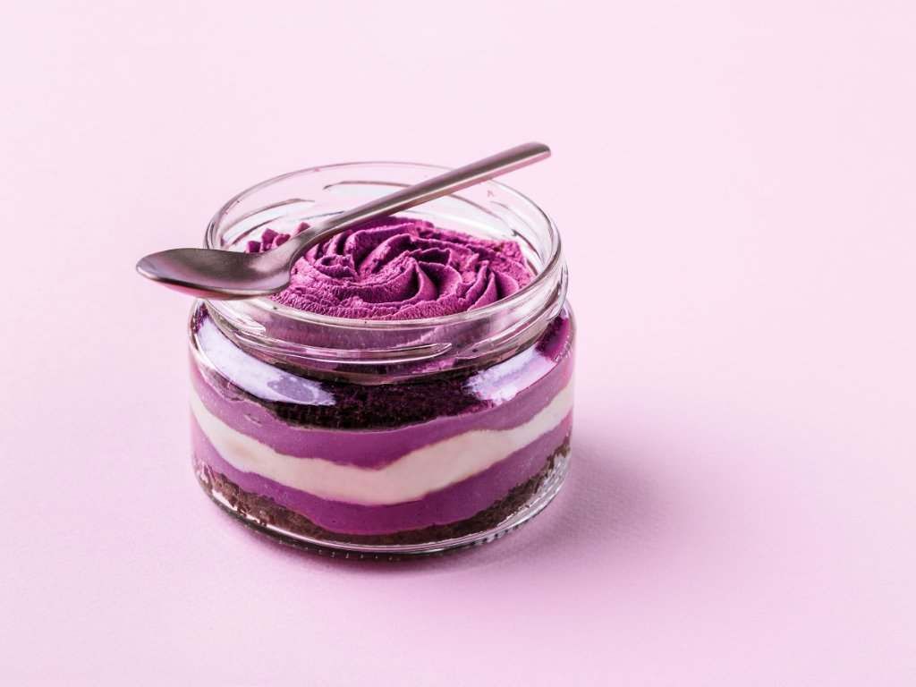 Un tarro de postre morado con una cuchara dentro sobre un fondo rosa.