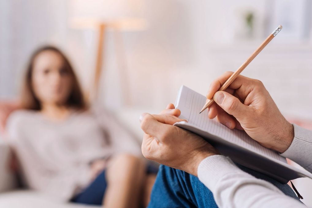 Gros plan sur une personne écrivant dans un carnet, avec une personne floue se reposant sur un canapé en arrière-plan.