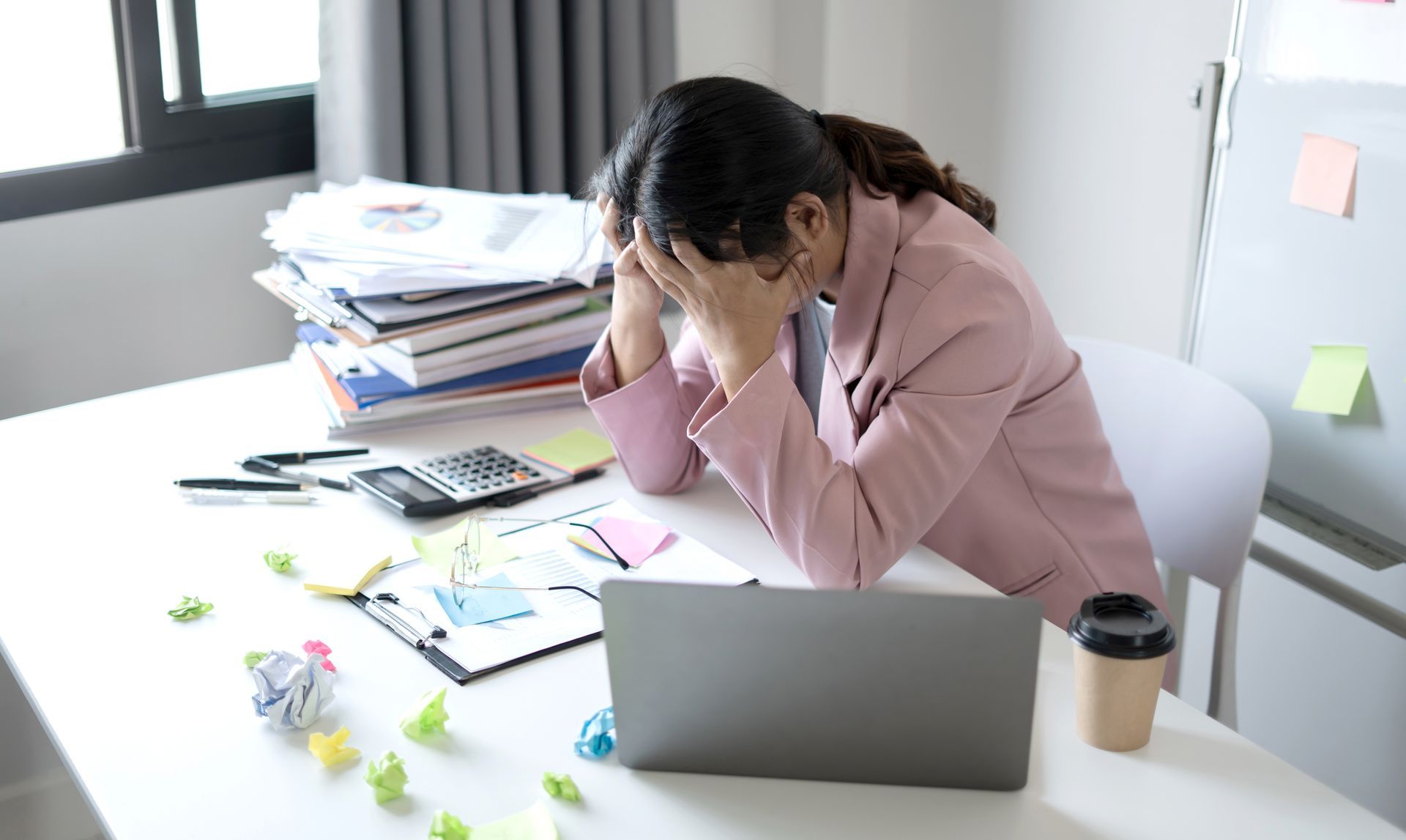 Une personne vêtue d'un blazer rose est assise à un bureau encombré, un ordinateur portable à la main, l'air stressé, la tête entre les mains.