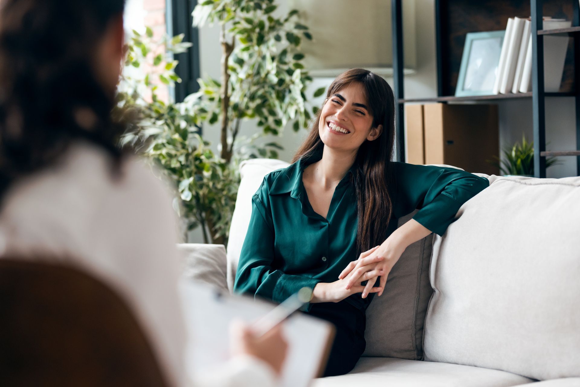 Une personne assise sur un canapé sourit à un thérapeute qui prend des notes lors d'une consultation dans un cabinet lumineux.