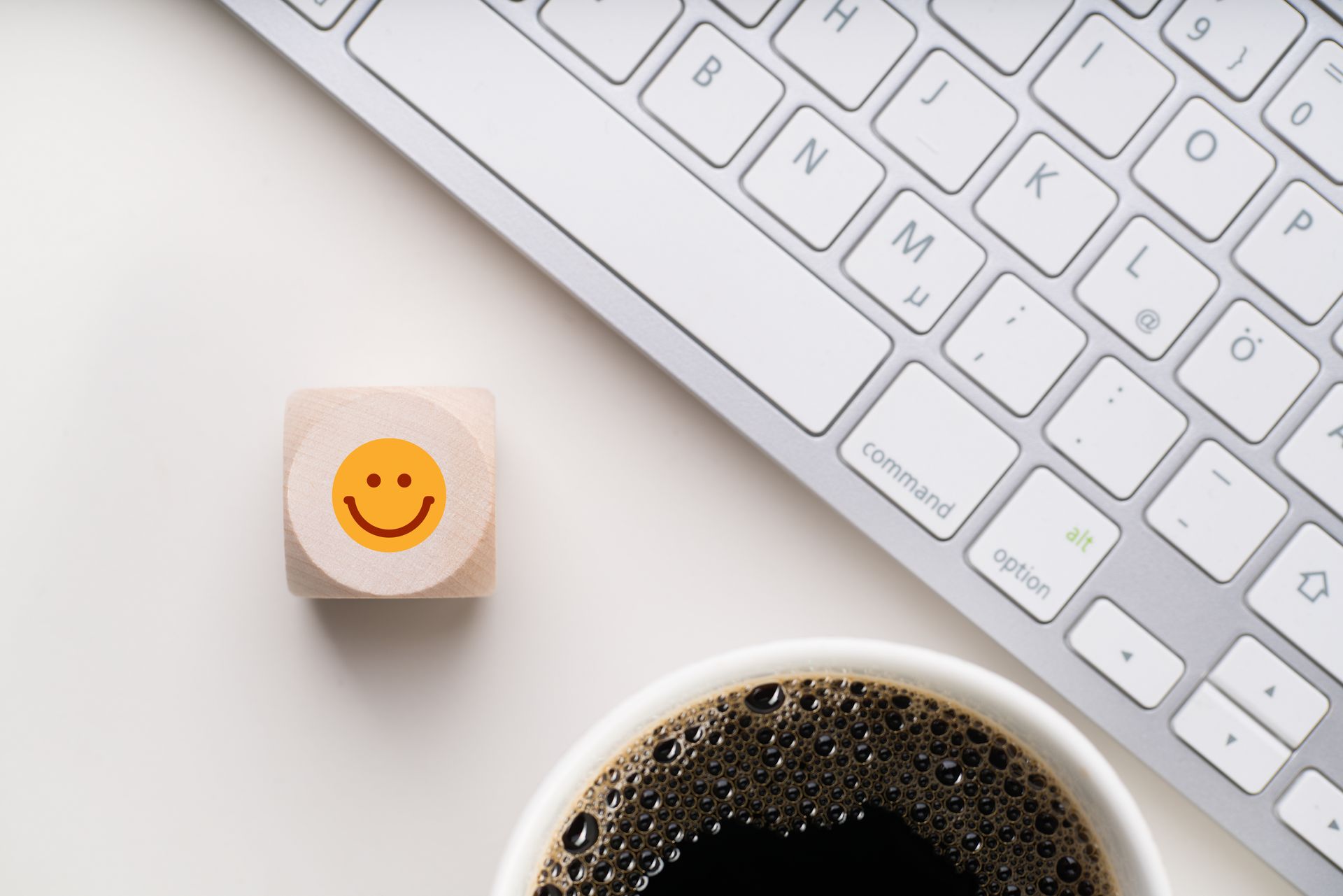 Un clavier blanc, une tasse de café noir et un bloc de bois orné d'un smiley jaune sur une surface blanche.