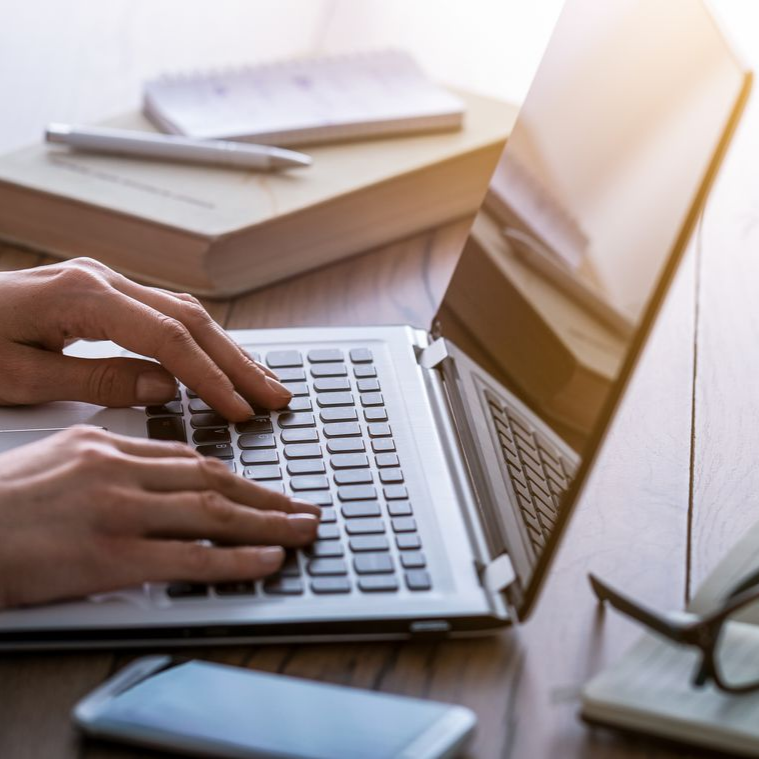 Des mains tapent sur un ordinateur portable argenté posé sur un bureau en bois, avec un carnet, un stylo et un smartphone à proximité.