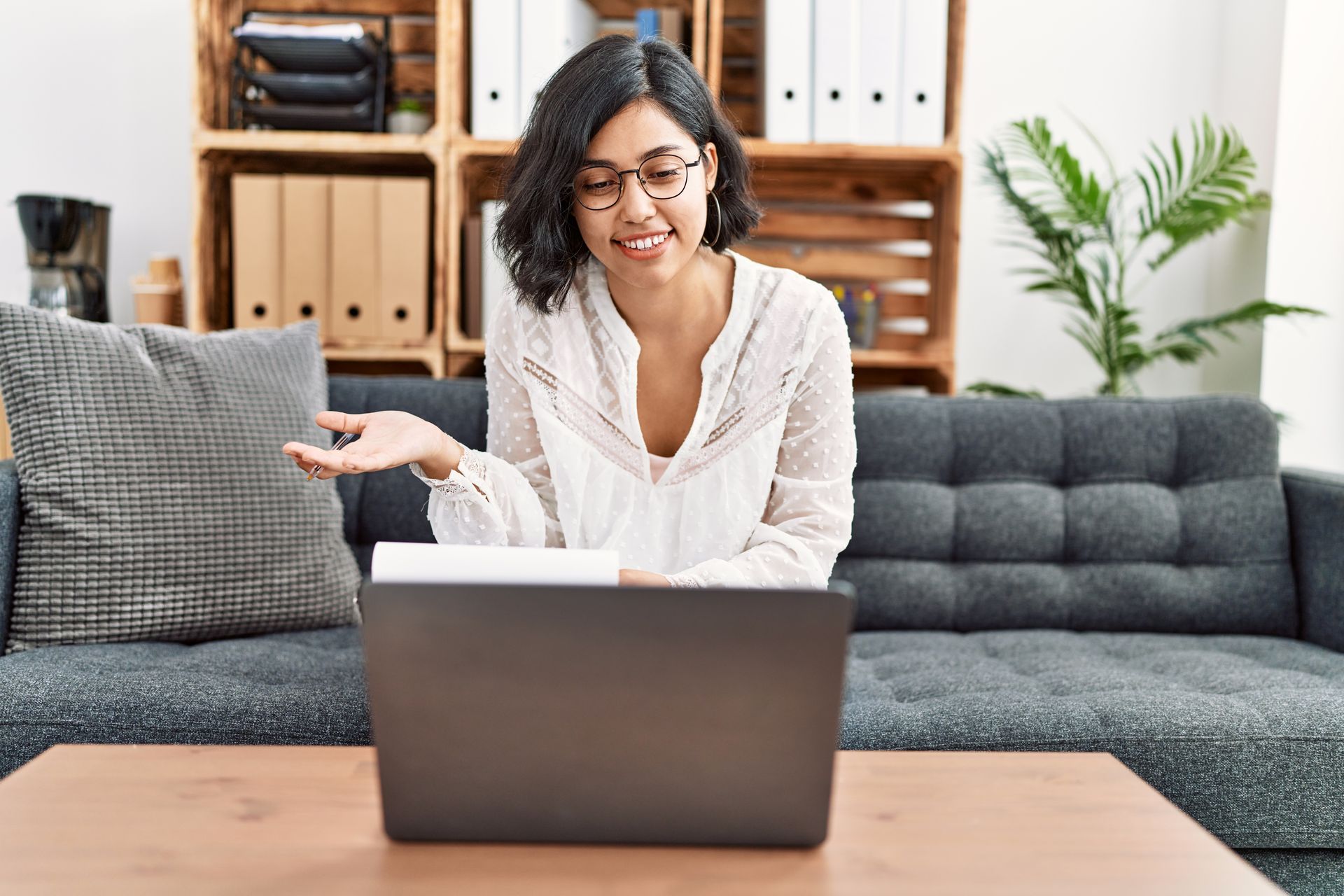 Dans un espace de bureau lumineux, une personne vêtue d'un haut blanc est assise sur un canapé, regardant un ordinateur portable et faisant un geste de la main.