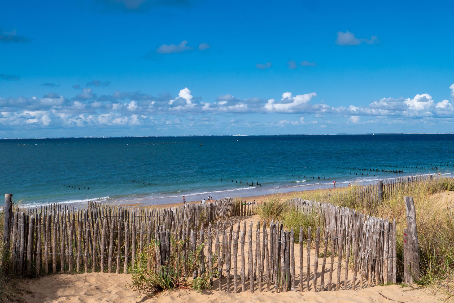 Une clôture rustique en bois mène à une plage de sable fin et à un océan bleu calme, sous un ciel lumineux parsemé de nuages blancs.