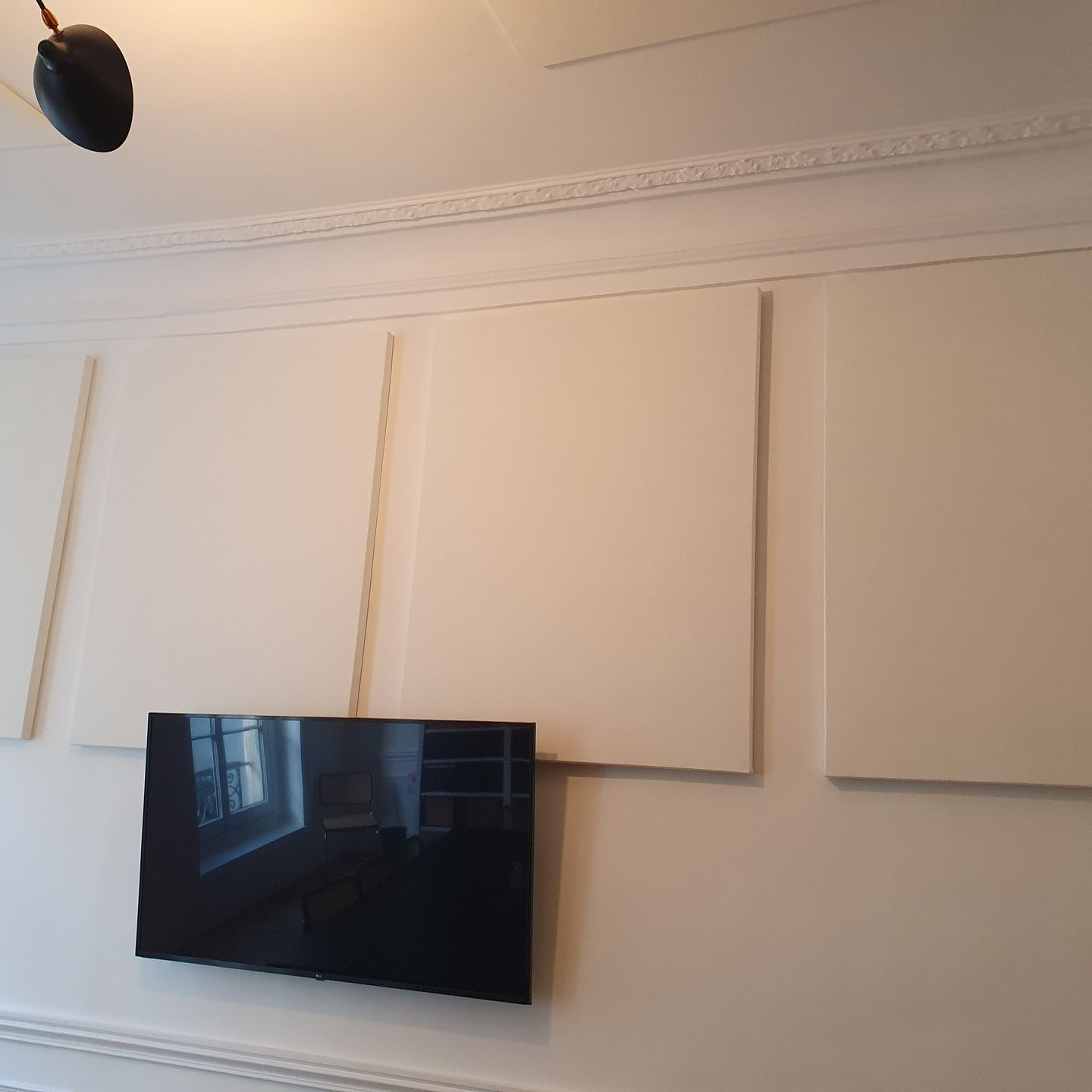 Téléviseur mural sous des panneaux acoustiques beiges sur un mur blanc dans une pièce.