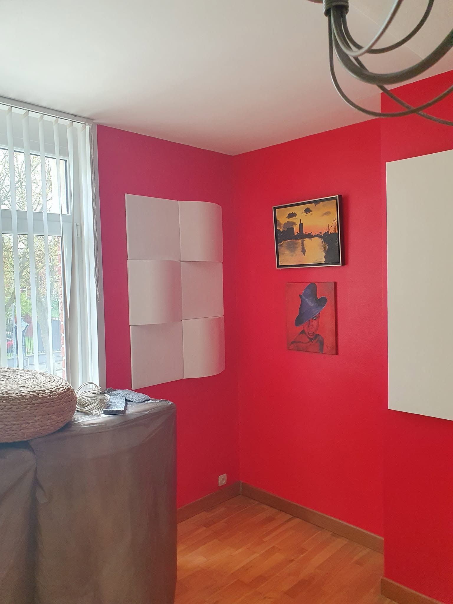 Pièce aux murs rouges avec des tableaux, des stores blancs et des meubles emballés près de la fenêtre.