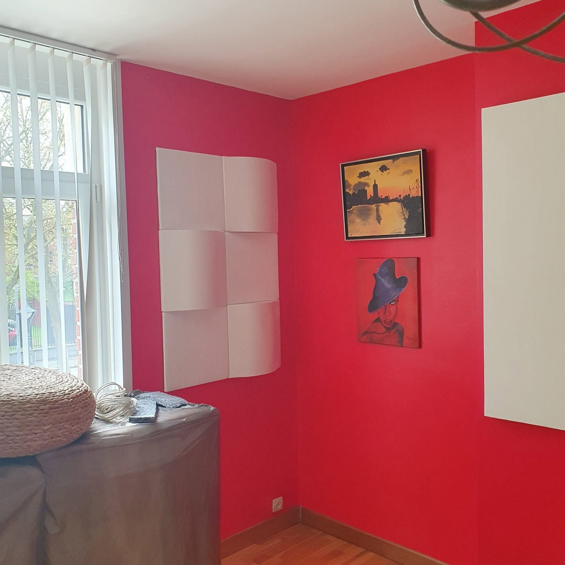 Pièce aux murs rouges avec des tableaux, des stores blancs et des meubles emballés près de la fenêtre.