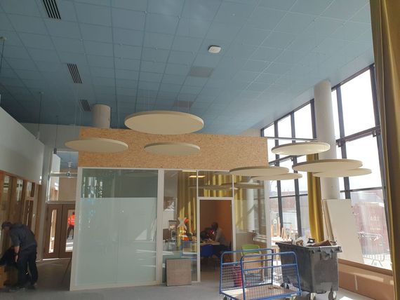 Hall d'entrée moderne avec baffles acoustiques suspendus au plafond, parois vitrées et une personne assise à l'intérieur.